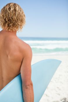 Young blonde man holding his blue surfboard while standing in front of the ocean
