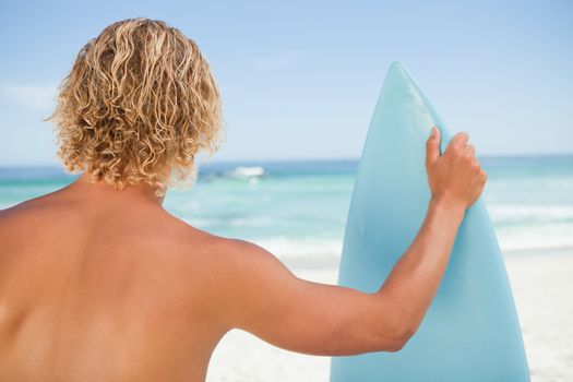 A young blonde man holding a perched surfboard while standing on the beach