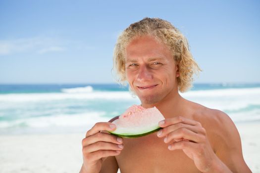 Smiling man holding a piece of a watermelon while standing in front of the ocean