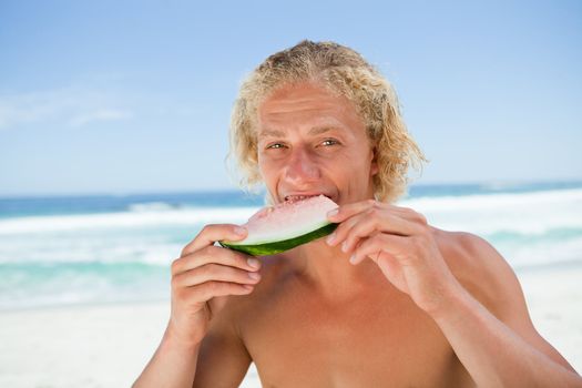 Young man eating a piece of watermelon while standing on the beach