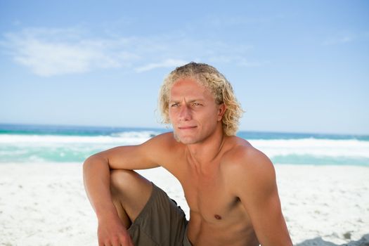 Young blonde man sitting on the beach while attentively looking towards the side
