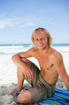 Smiling blonde man sitting on his blue beach towel while looking in front of him