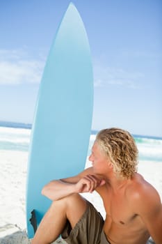 Serious blonde man sitting in front of the sea next to his blue surfboard 