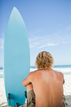 Blonde man sitting down with his arms around his legs next to his surfboard