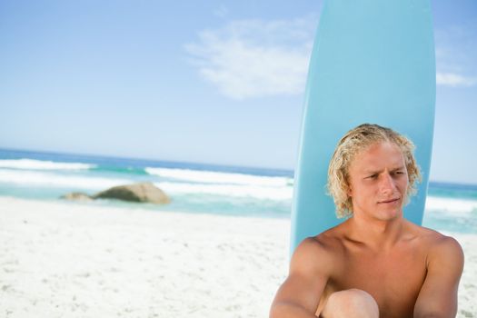 Blonde man sitting on the beach with his surfboard while looking towards the side