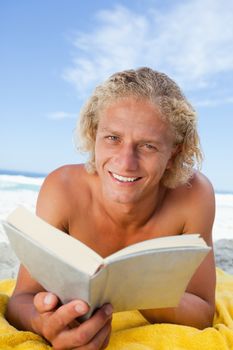 Smiling blonde man reading a book while lying on his yellow beach towel