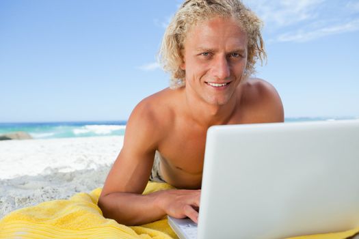 Blonde man lying on the beach while using his laptop and showing a great smile