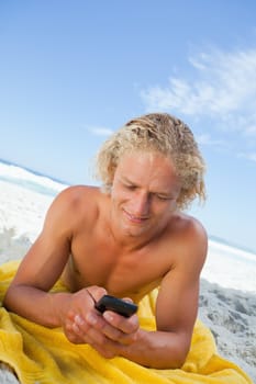 Blonde man lying on the beach while sending a text with his cellphone
