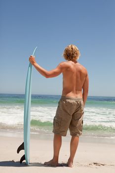 Young blonde man waiting for waves while holding his surfboard in front of the ocean