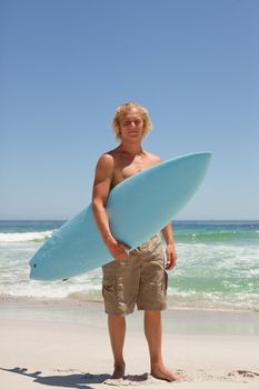 Blonde man holding his blue surfboard while standing on the beach