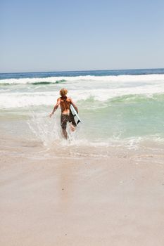 Young blonde man running in the water while holding his blue surfboard
