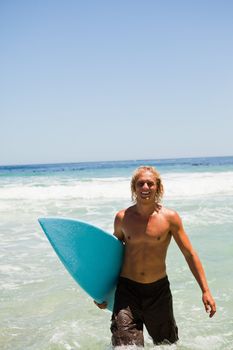 Blonde man walking in the water while holding his surfboard and smiling