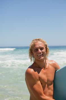 Young blonde man looking at the camera while smiling and holding his surfboard