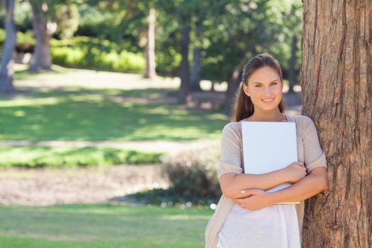 Smiling young woman with a laptop leaning against a tree