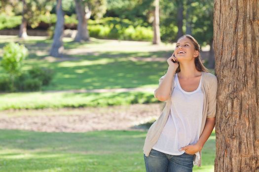 Cheerful young woman on the phone leaning against a tree