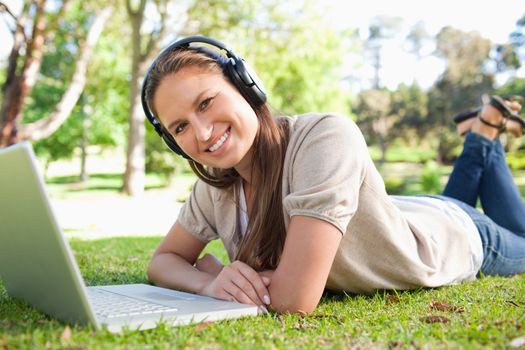 Smiling young woman with a headset and a laptop lying on the lawn
