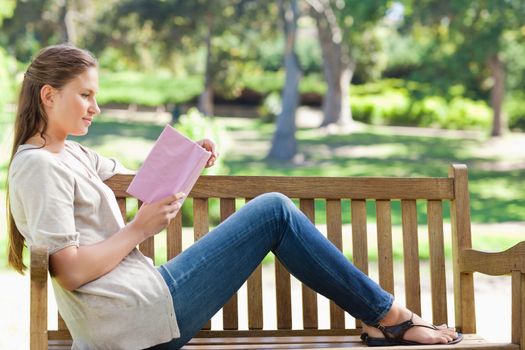 Side view of a young woman reading a novel on a park bench