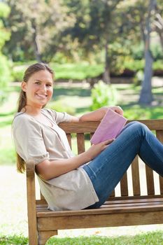 Side view of a smiling young woman with a book on a park bench