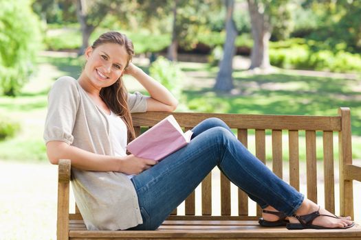 Side view of a smiling young woman sitting on a park bench with her book