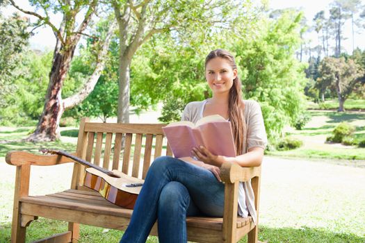 Smiling young woman with a book and a guitar sitting on a bench