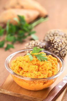 Sweet potato spread in glass bowl garnished with parsley with small sesame-wholegrain buns on wooden board (Selective Focus, Focus on the front of the parsley leaf on the spread)