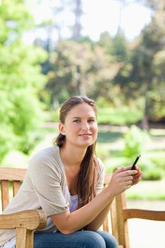 Smiling young woman holding her cellphone while on a park bench