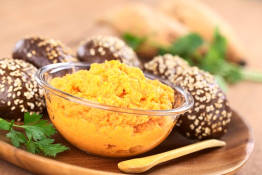 Sweet potato spread in glass bowl with small sesame-wholegrain buns, parsley and wooden spoon on wooden plate (Selective Focus, Focus one third into the spread)