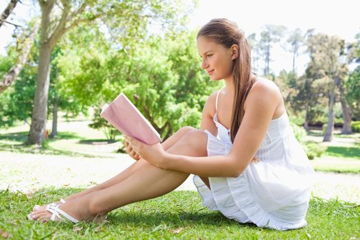 Side view of a young woman sitting on the lawn while reading a book