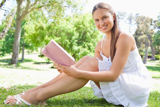 Side view of a smiling young woman sitting on the lawn with a book