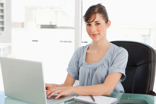 An attractive employee sits at her desk while typing on her laptop and looking into the camera