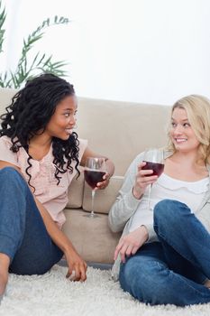 Two women are talking and sitting up against a couch drinking wine