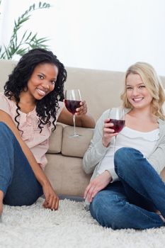 Two women sitting on the ground holding wine glasses are smiling at the camera