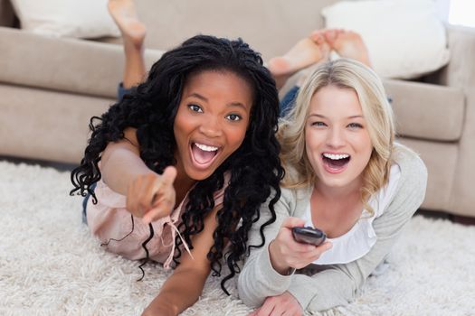 Two laughing women are lying on the ground and looking at the camera with a TV remote control