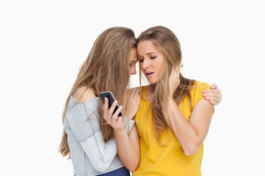 Upset young woman looking her cellphone consolded by her friend against white background