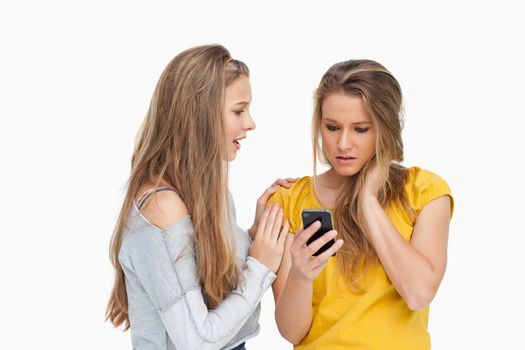 Upset young woman holding her cellphone consolded by her friend against white background