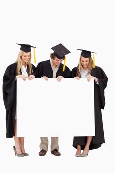 Three smiling students in graduate robe holding a blank sign against white background