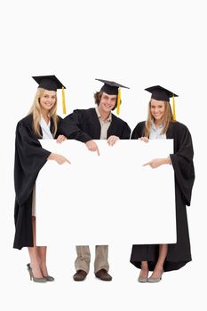 Three students in graduate robe holding and pointing a blank sign against white background