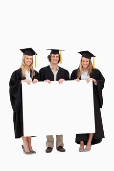 Three students in graduate robe holding a blank sign against white background