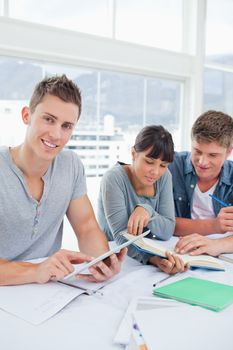 A smiling student using a tablet looks into the camera as his friends use a book to find the answer