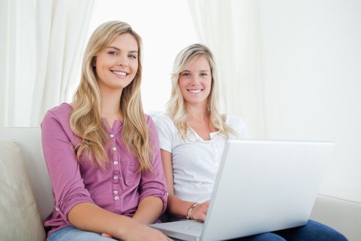 Sisters sitting and smiling on the couch looking at the camera as they hold a laptop