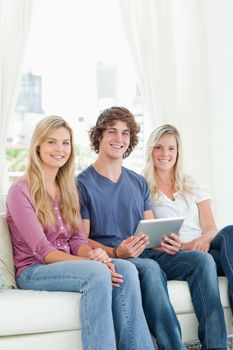 Two sisters and a brother sit together on the couch as the brother holds the tablet while they all look at the camera 