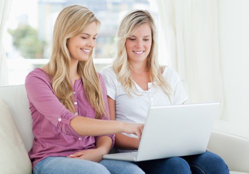 Two smiling women using a laptop as one points to the screen while they sit on the couch together