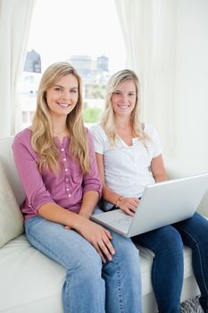 Sisters smiling as they sit on the couch together and hold a laptop while looking into the camera