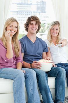 Three friends eating popcorn while smiling and using the tv remote to change the channel 