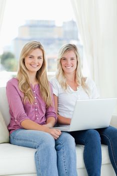 A pair of sisters sit together with a laptop on the couch as they look at the camera 