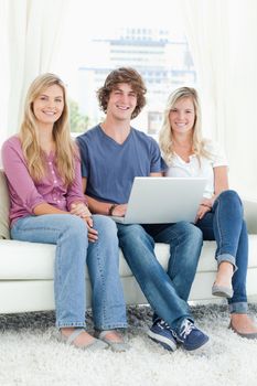 A smiling group of friends sit together while looking at the camera with a laptop in their hands 