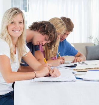 A girls sits at the table smiling and looking at the camera while her friends study