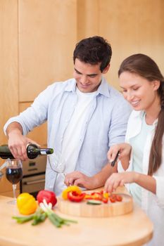 Portrait of a young man pouring a glass of wine while his wife is cooking in their kitchen