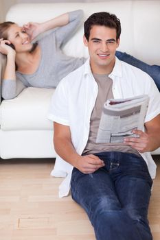Young man leaning against the sofa with newspaper while his girlfriend listens to music