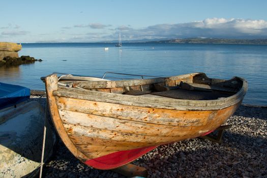 A wooden boat out of water on a pebble beach with the sea and blue sky in the distance